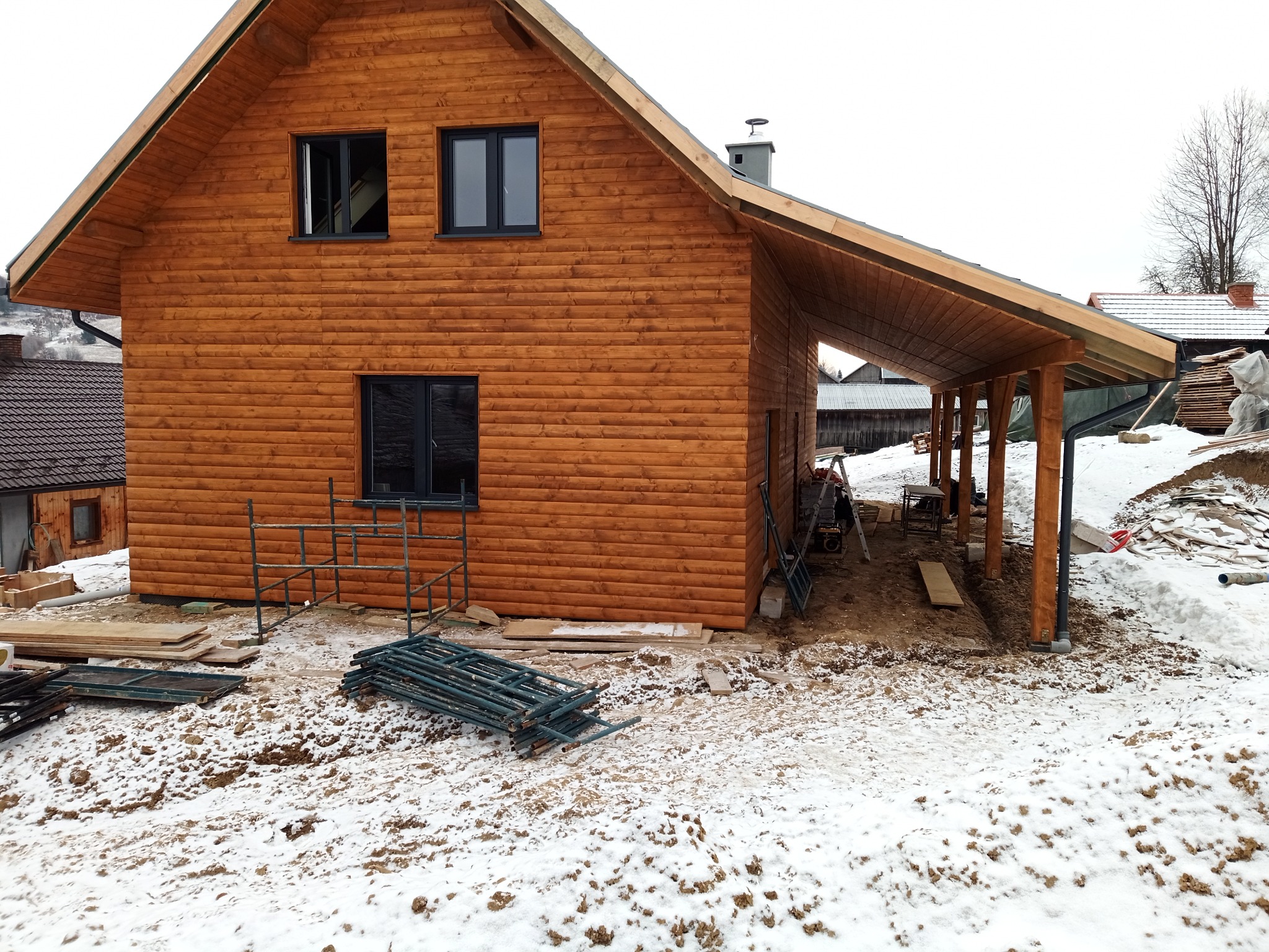 Surowy drewniany dom z ciemnymi oknami w zimowej scenerii, rusztowania leżące na śniegu, widoczny fragment zadaszenia i przygotowane deski.