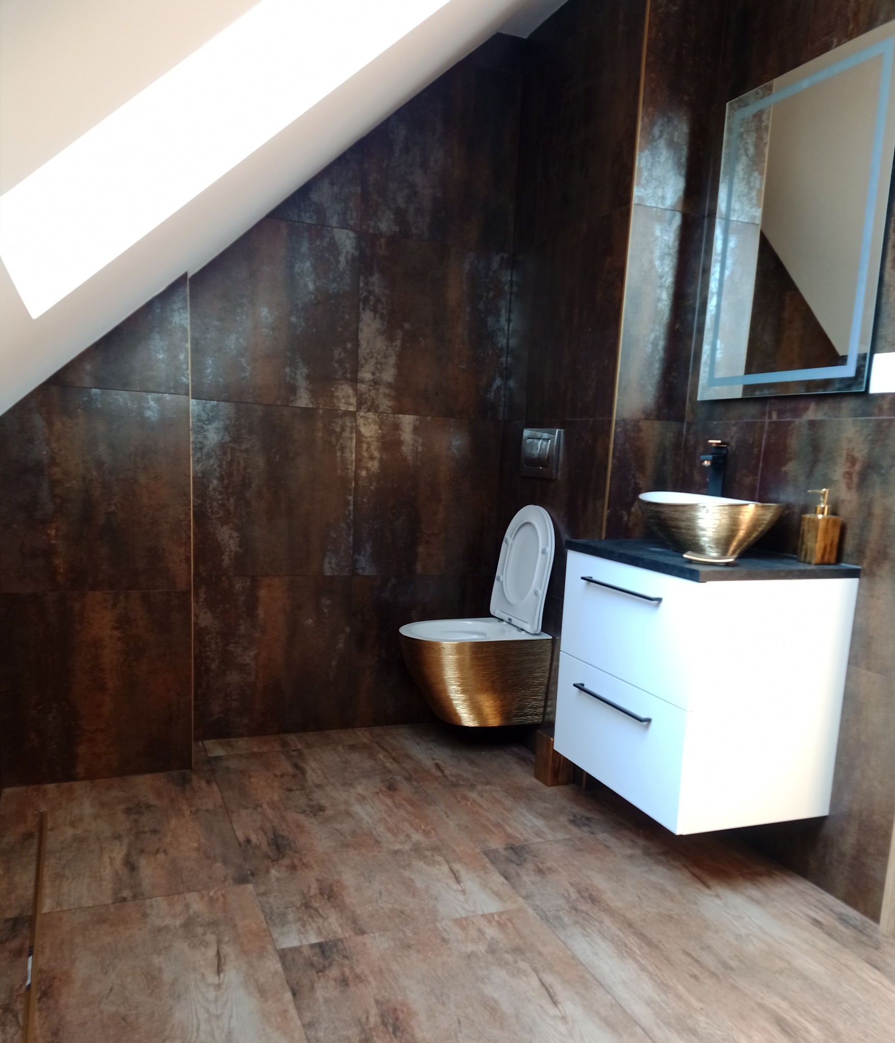 Nowoczesna łazienka na poddaszu z brązowymi płytkami imitującymi rdzę, złotą miską umywalkową i toaletą w tym samym kolorze, białą szafką pod umywalką z czarnymi uchwytami oraz podświetlanym lustrem.