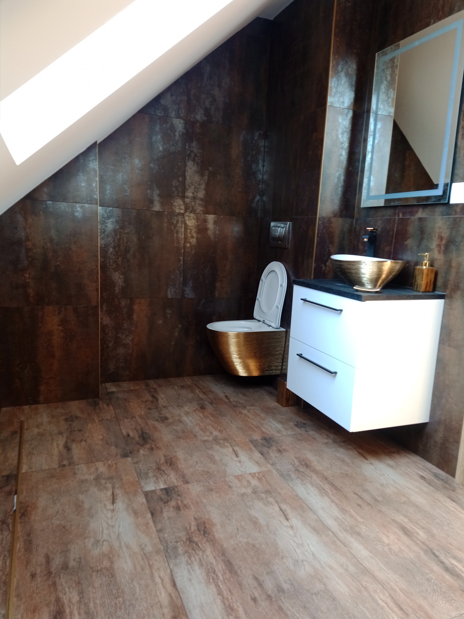 Nowoczesna łazienka na poddaszu z brązowymi płytkami imitującymi rdzę na ścianach, drewnopodobna podłoga, złota umywalka i toaleta, biała szafka z czarnym blatem i podświetlane lustro.