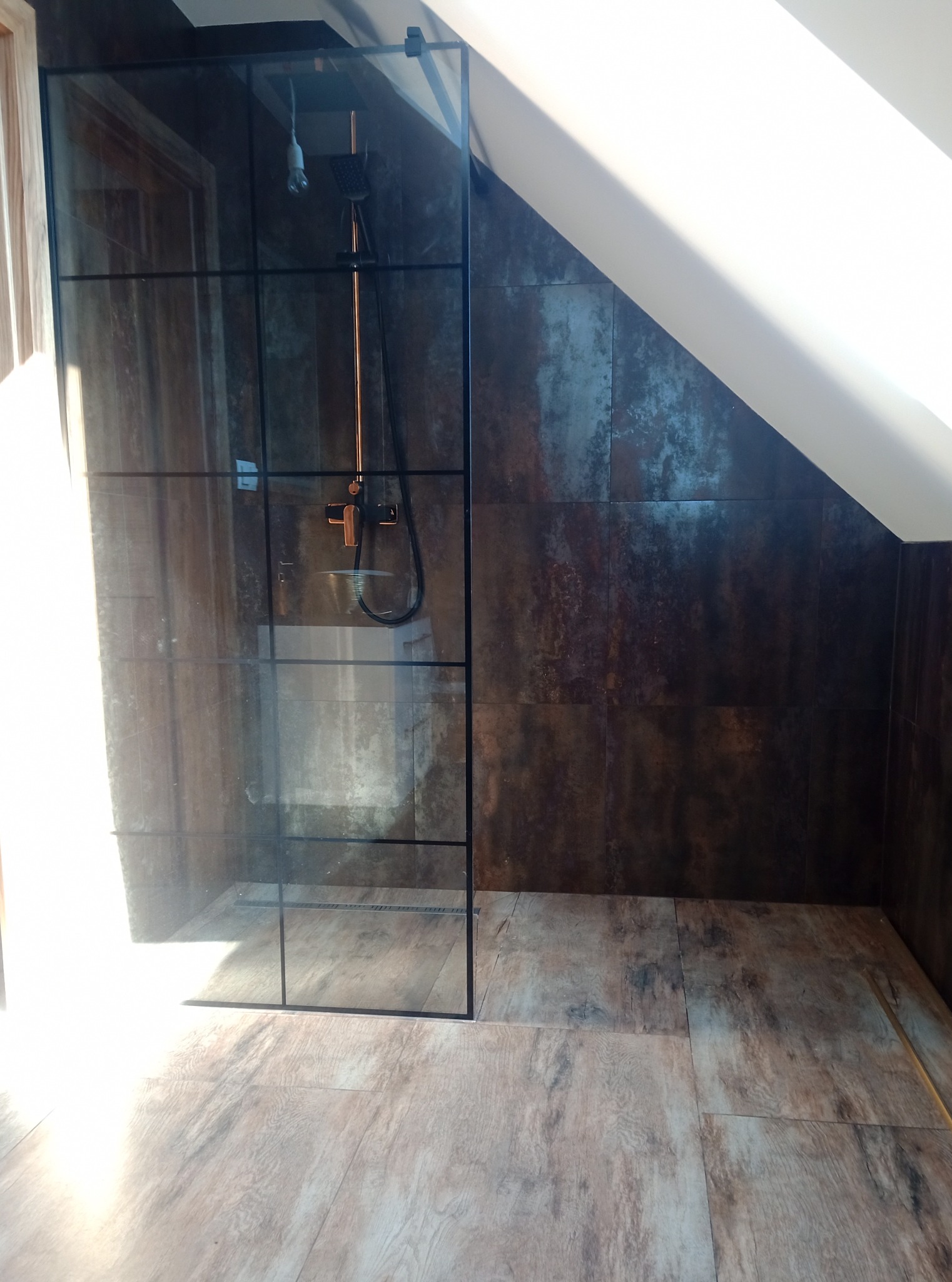 Nowoczesna kabina prysznicowa z czarnymi profilami w łazience na poddaszu, ściany wyłożone płytkami imitującymi rdzę, podłoga drewnopodobna.