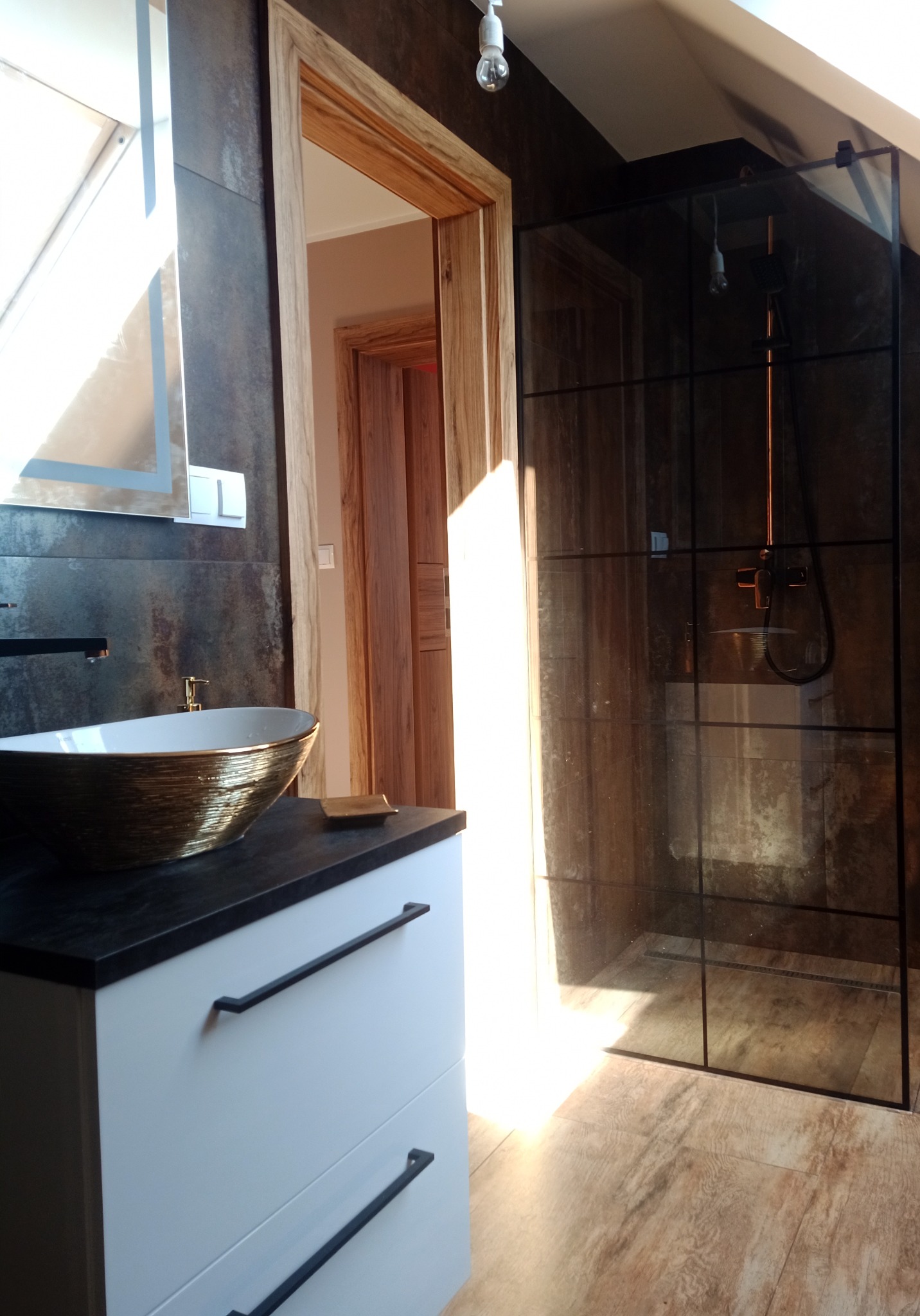 Nowoczesna łazienka z kabiną prysznicową typu walk-in z czarnymi profilami, umywalką nablatową w kolorze białym ze złotymi detalami i szafką z białymi frontami i czarnymi uchwytami, widok...