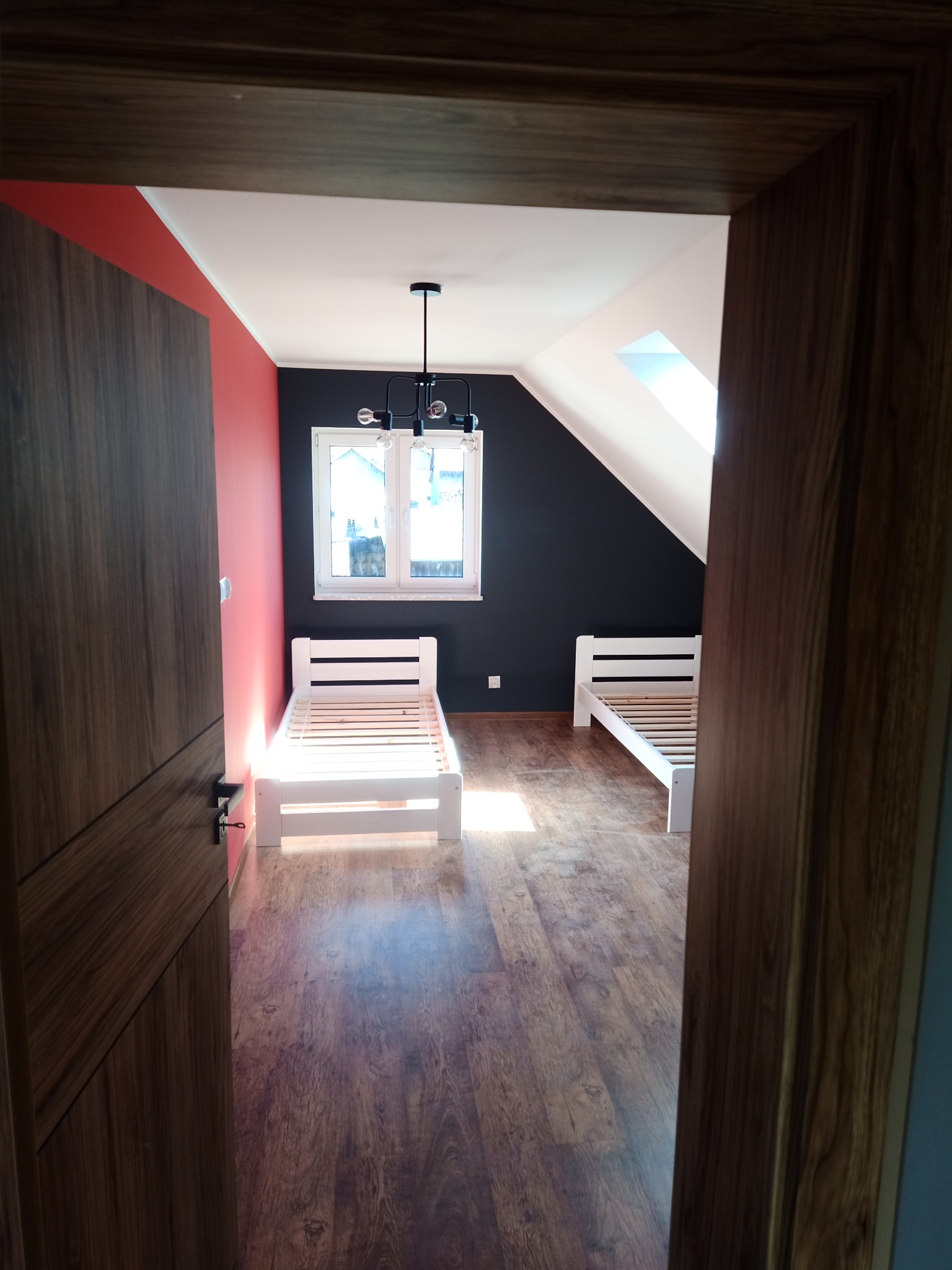 Pokój na poddaszu z drewnianą podłogą, dwoma białymi łóżkami z drewnianymi stelażami, oknem dachowym i lampą sufitową w stylu industrialnym, widok zza drewnianej framugi drzwi.