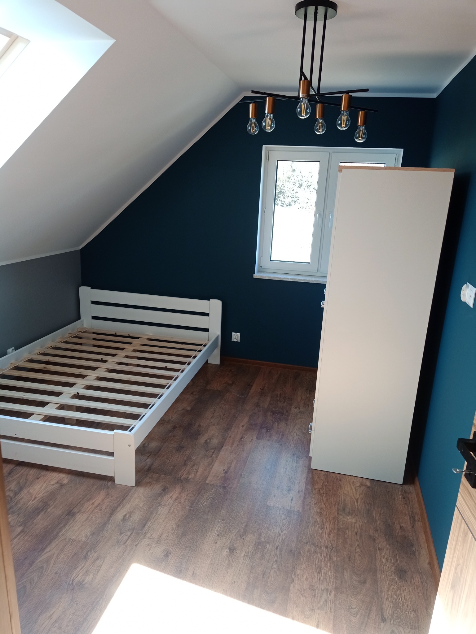 Poddasze z drewnianą podłogą, białym łóżkiem bez materaca, białą szafą i nowoczesną lampą wiszącą na tle ciemnoniebieskiej ściany.
