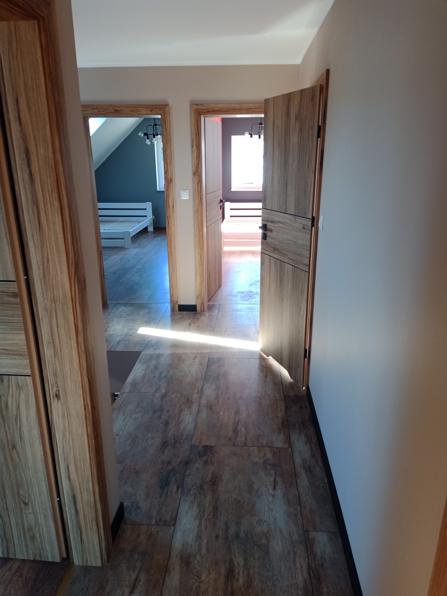 Wnętrze domu z otwartymi drewnianymi drzwiami, widok na podłogę z paneli imitujących drewno i dalsze pomieszczenia, oświetlone naturalnym światłem.