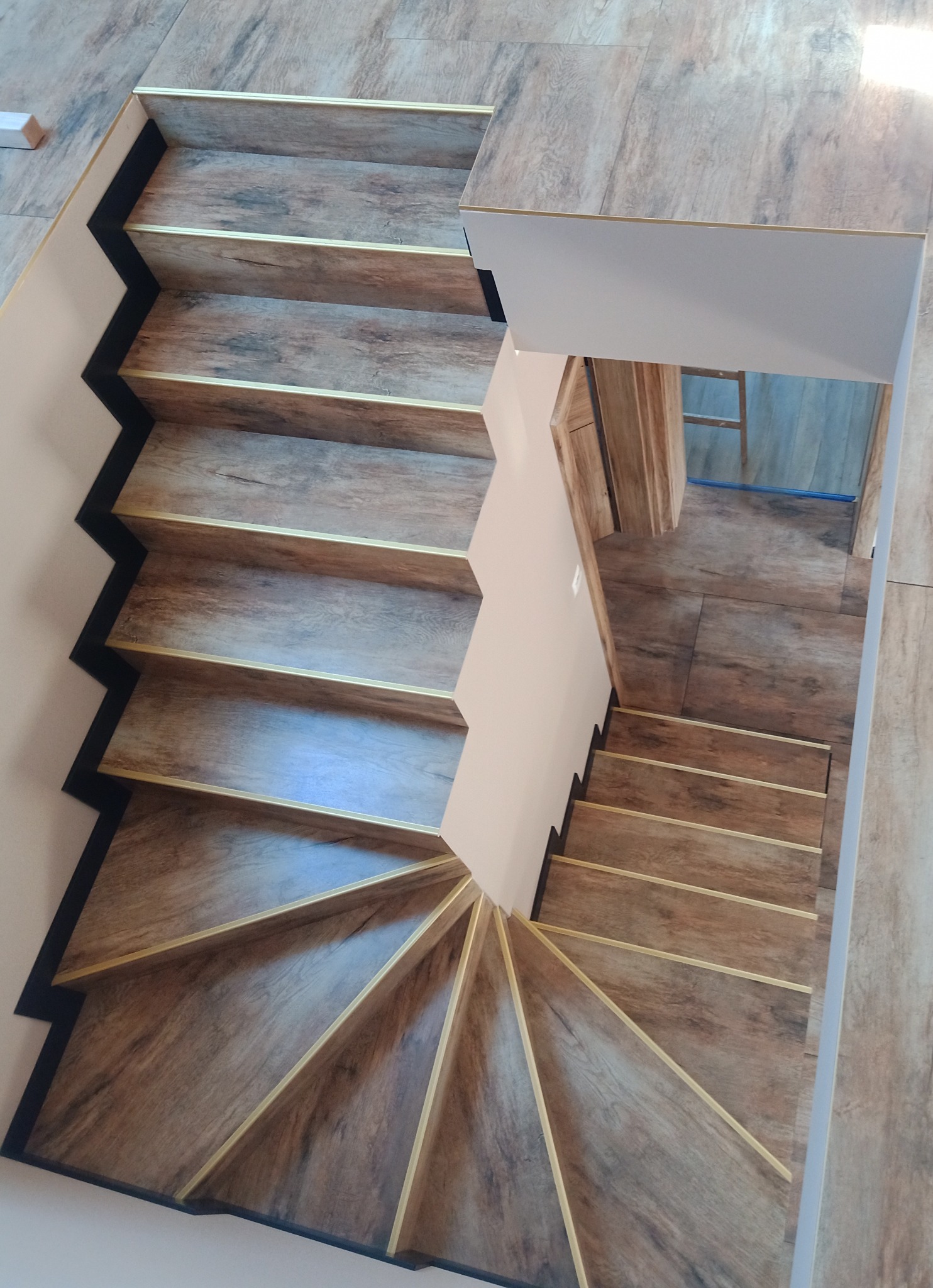 Spiralne schody wykończone panelami drewnopodobnymi z subtelnymi złotymi listwami, widok z góry, krawędzie pomalowane na czarno.