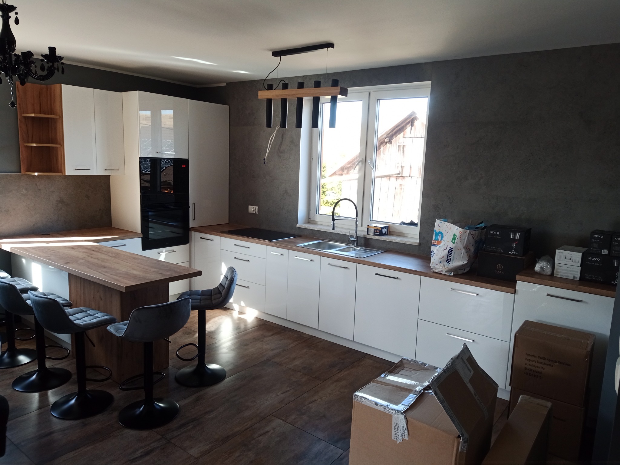 Nowoczesna kuchnia z wyspą i hokerami, białe szafki, ciemnoszara ściana, drewniany blat, kartony z akcesoriami na podłodze, widok na zewnątrz przez okno.