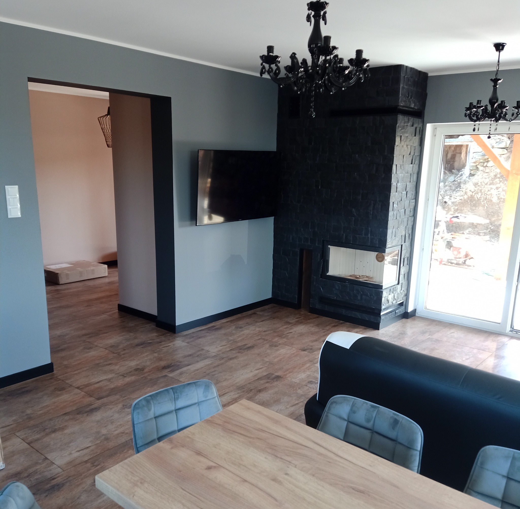 Szare wnętrze domu z czarnym kominkiem wyłożonym kamieniem, drewnianą podłogą i stołem z krzesłami, widok na sąsiednie pomieszczenie przez czarną ramę drzwiową.