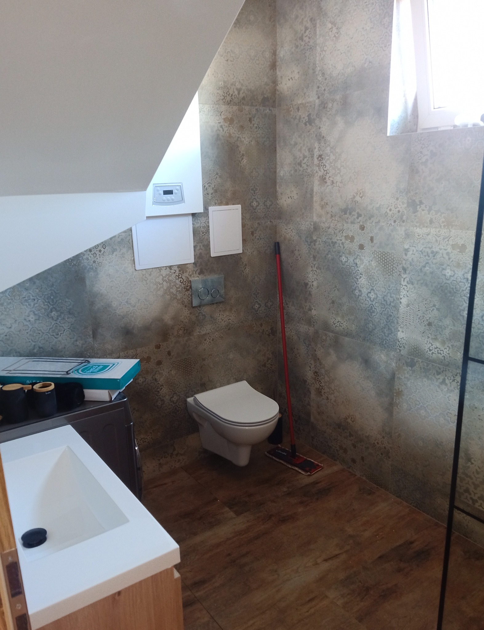 Wykończona łazienka na poddaszu z umywalką nablatową, toaletą wiszącą i wzorzystymi płytkami ceramicznymi na ścianach, widoczny bojler oraz mop.