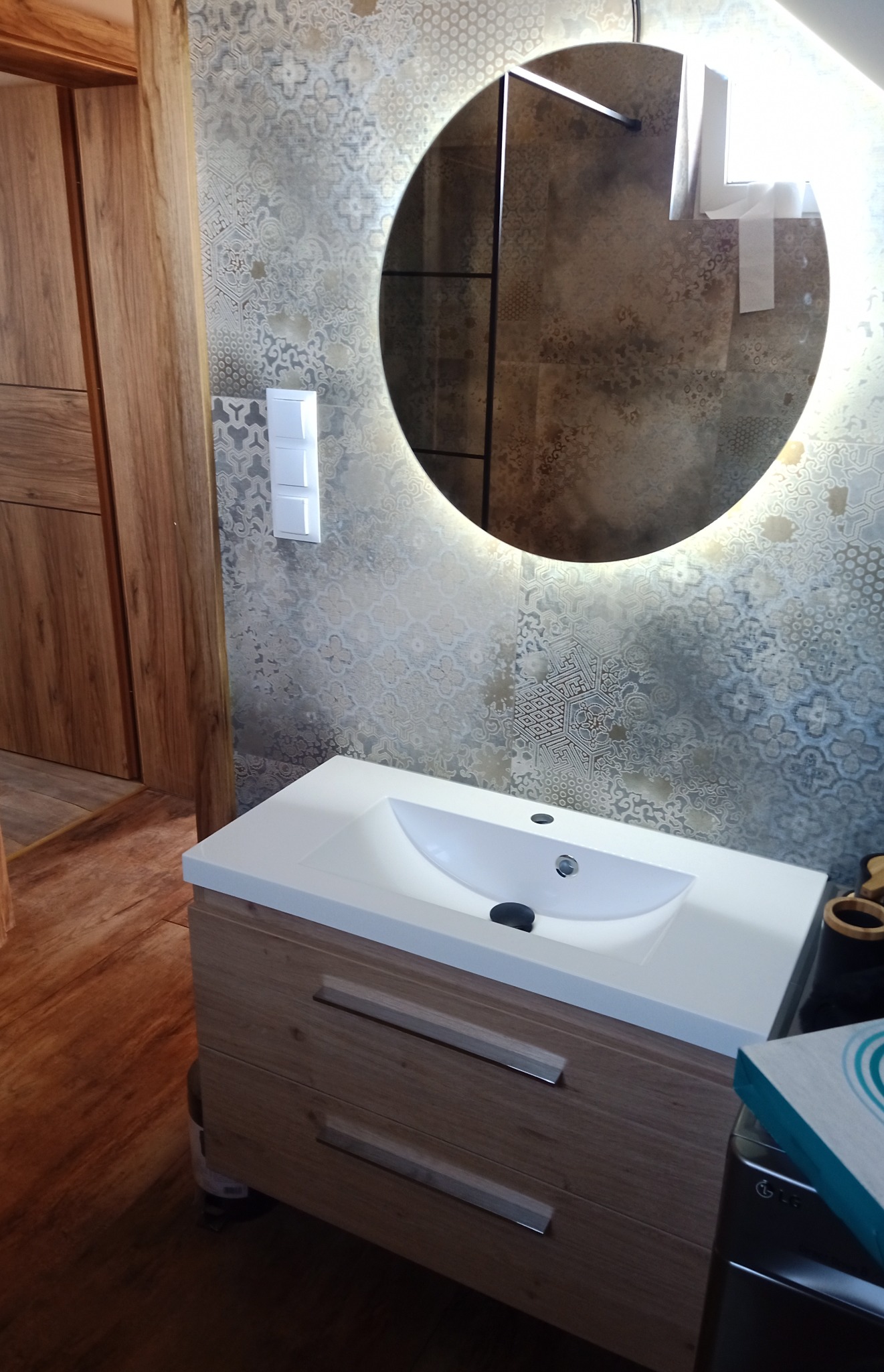 Nowoczesna łazienka z umywalką nablatową na szafce z drewnopodobnymi frontami, okrągłe lustro z podświetleniem LED i wzorzyste płytki na ścianie.
