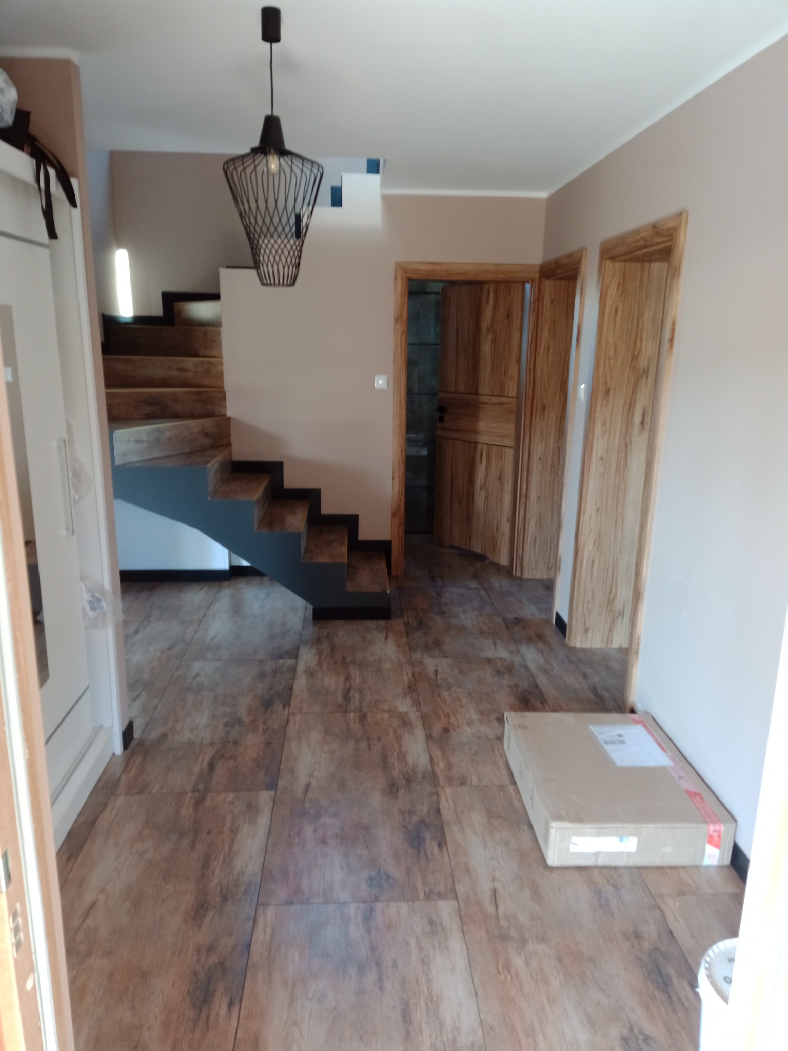 Wnętrze domu w trakcie wykończenia: schody drewniane z szarymi elementami, podłoga z płytek imitujących drewno, dwie drewniane drzwi oraz karton na podłodze.