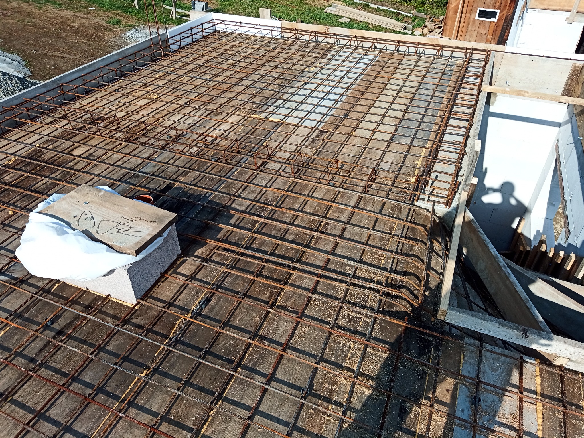 Zbrojenie stropu budynku w trakcie budowy, widoczne pręty zbrojeniowe ułożone w kratę, szalunek z białego styropianu na obrzeżach, cień osoby fotografującej.