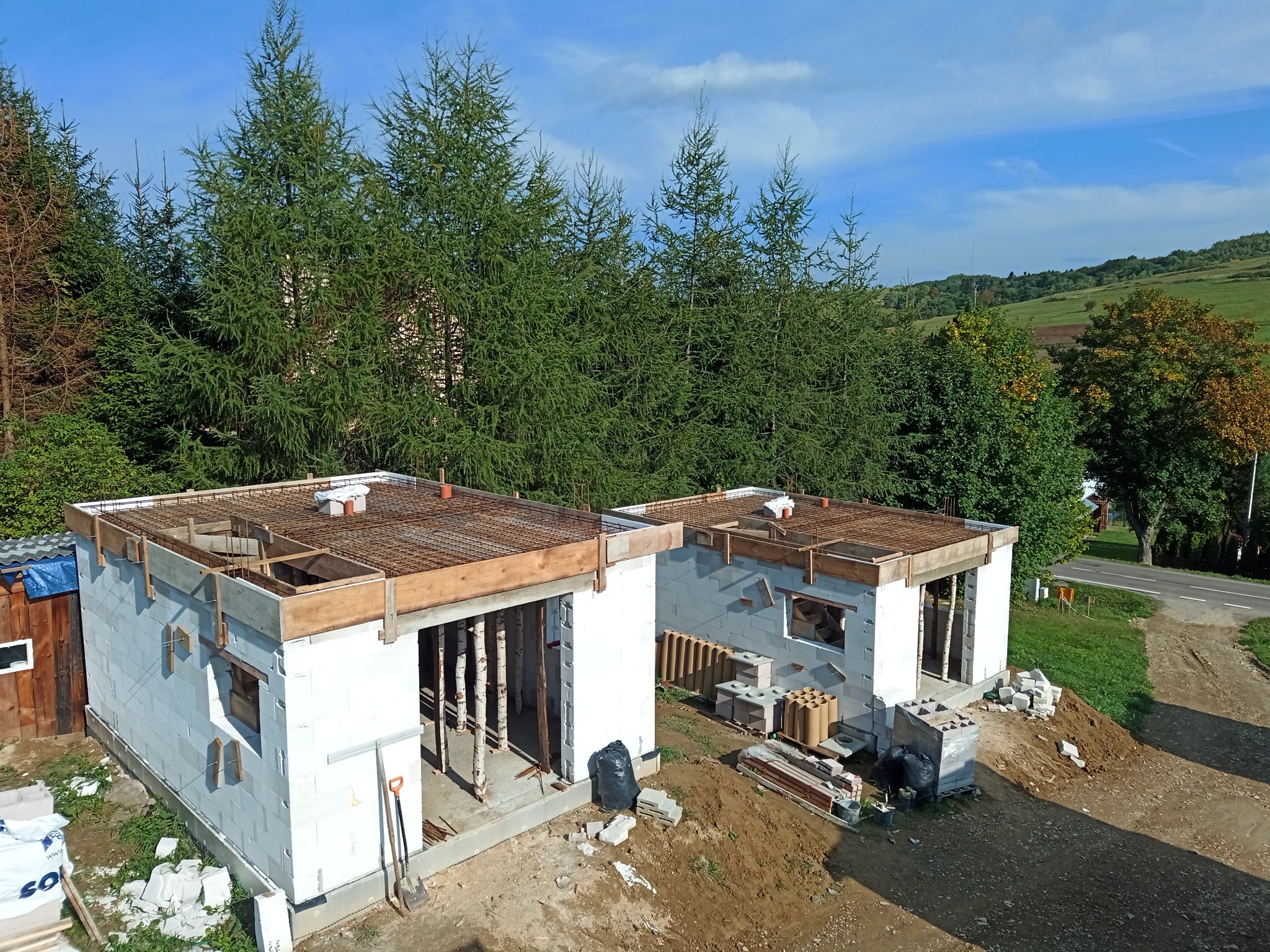 Dwa surowe domy murowane w stanie surowym otwartym, widoczne z góry, zbrojenie stropu, białe bloczki, podpory z bali, okolica zalesiona i pagórkowata w tle.
