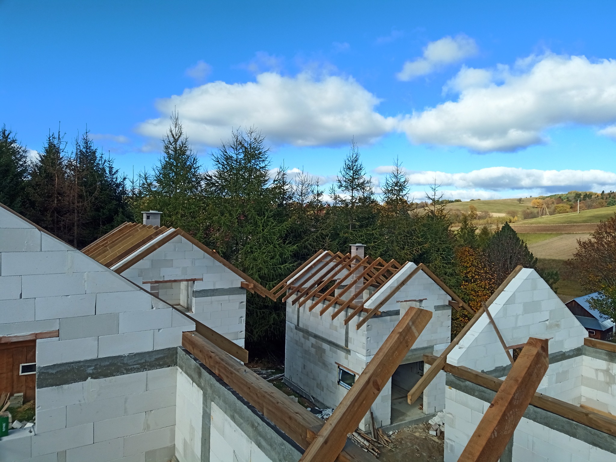 Wysoki kąt widzenia na kilka domów w trakcie budowy z białych bloczków, widać drewniane elementy konstrukcji dachu, w tle las iglasty i błękitne niebo z chmurami.