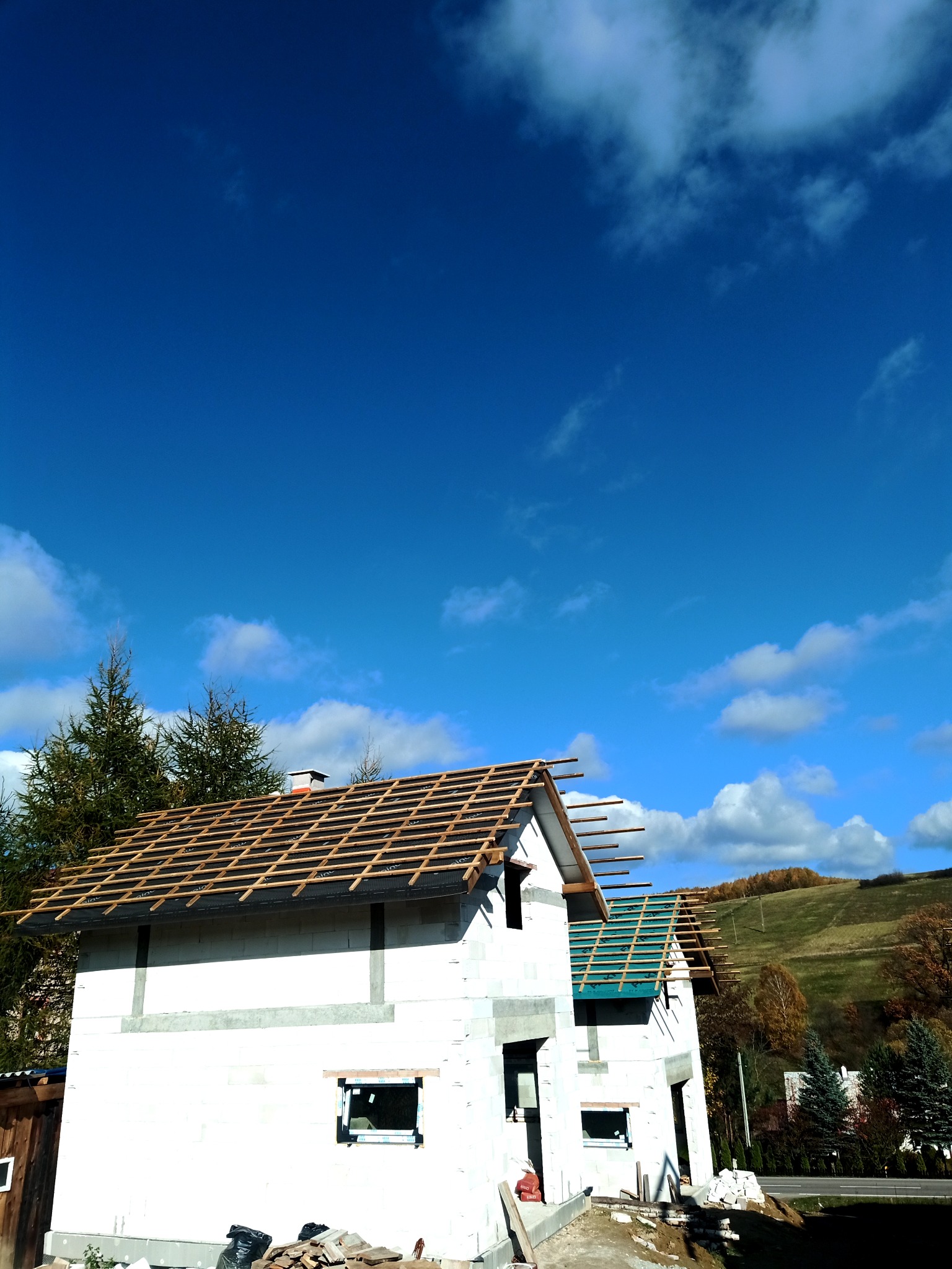 Surowy stan domu z białych bloczków, widoczna konstrukcja dachu z łatami, membrana dachowa częściowo położona, tło: błękitne niebo i wzgórza.