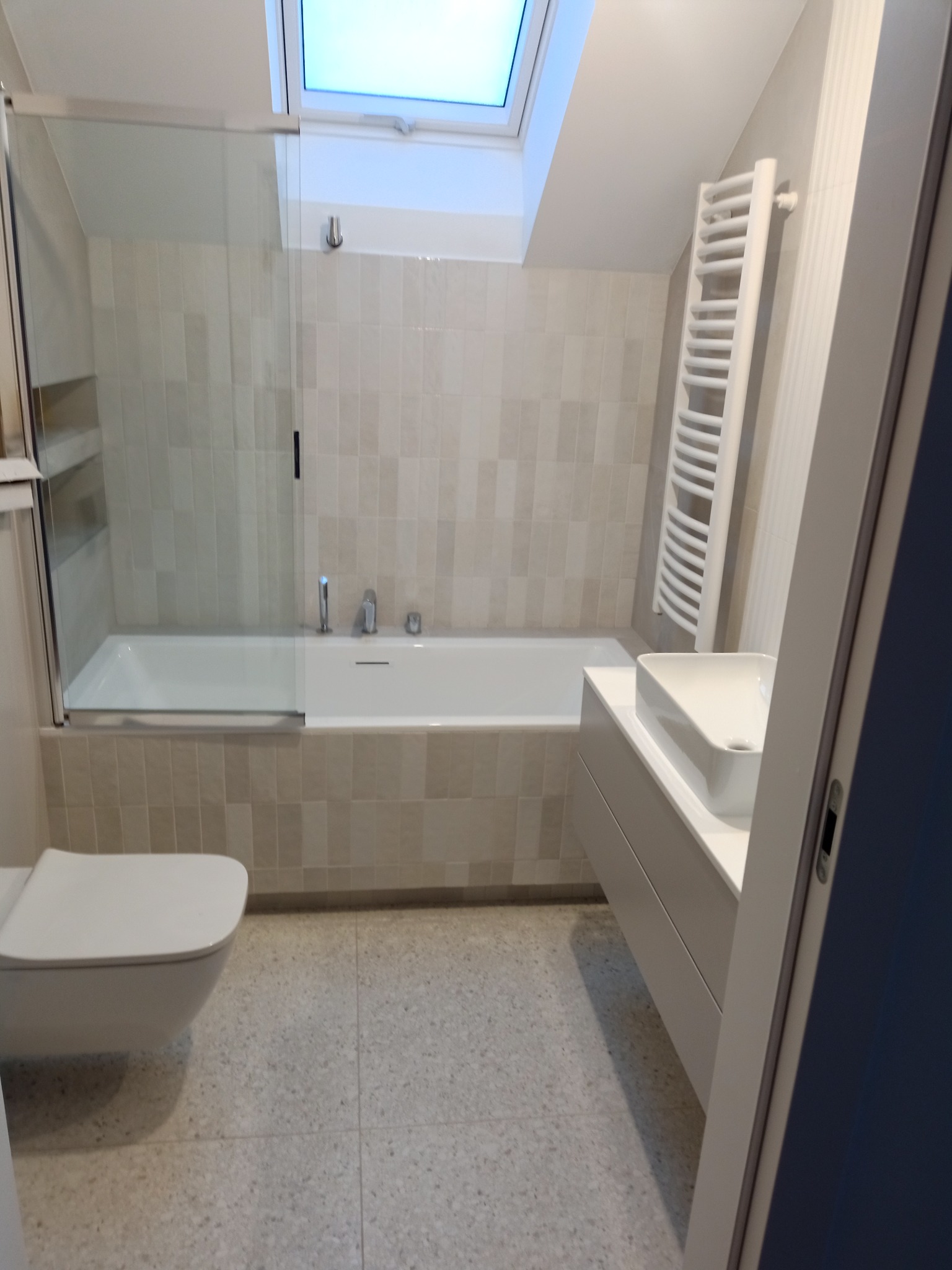 Nowoczesna łazienka z beżowymi płytkami, białą wanną z szklanym parawanem, wiszącą toaletą i grzejnikiem drabinkowym, oświetlona światłem dziennym z okna dachowego.