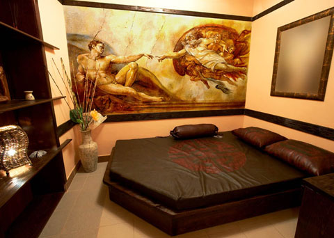 Pokój z łóżkiem i muralem nawiązującym do 'Stworzenia Adama' Michała Anioła, wazon z suchymi gałązkami po lewej, lustro na ścianie po prawej.