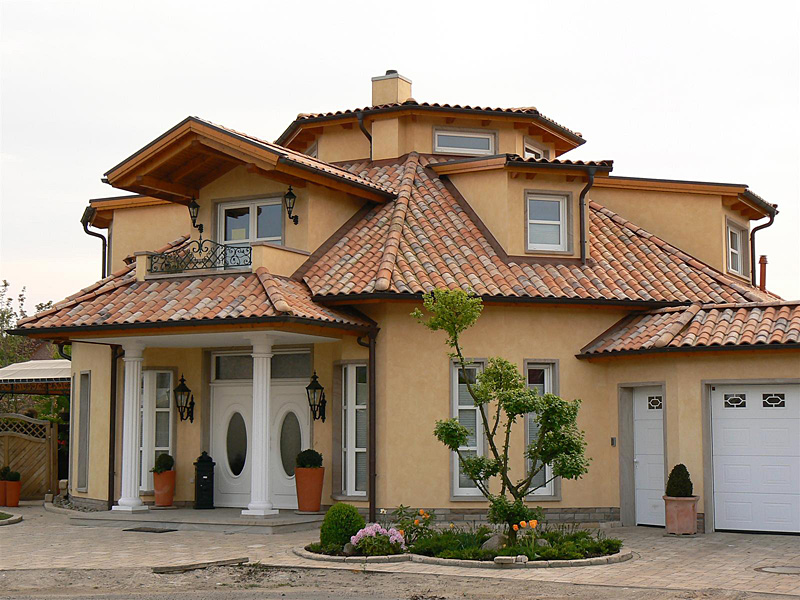 Widok z zewnątrz na dom w stylu śródziemnomorskim z dachem pokrytym ceramiczną dachówką w odcieniach czerwieni i brązu, jasną elewacją i kolumnami przy wejściu.