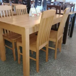 Jasny, drewniany stół z czterema krzesłami w sklepie meblowym, z widocznymi cenami na kartkach i innymi meblami w tle.