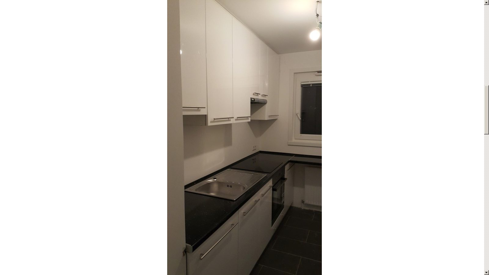 Biała, błyszcząca zabudowa kuchenna z czarnym blatem, widok na zlew i płytę grzewczą, ciemna podłoga z płytek, okno w tle.