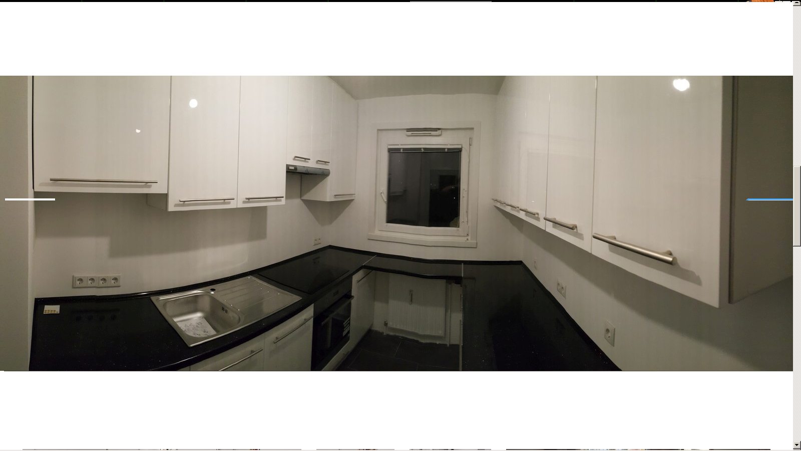 Nowoczesna kuchnia z białymi, błyszczącymi szafkami i czarnym blatem, widok na okno i elementy wykończenia.