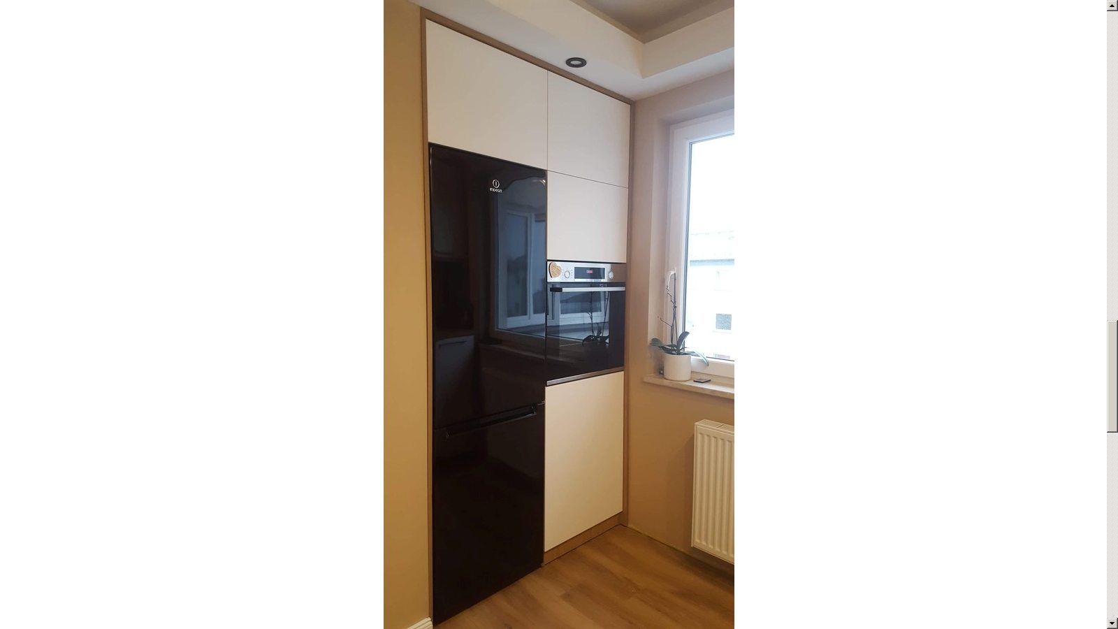 Nowoczesna zabudowa kuchenna z czarną lodówką i piekarnikiem w białych szafkach, widok na okno z parapetem i rośliną doniczkową.