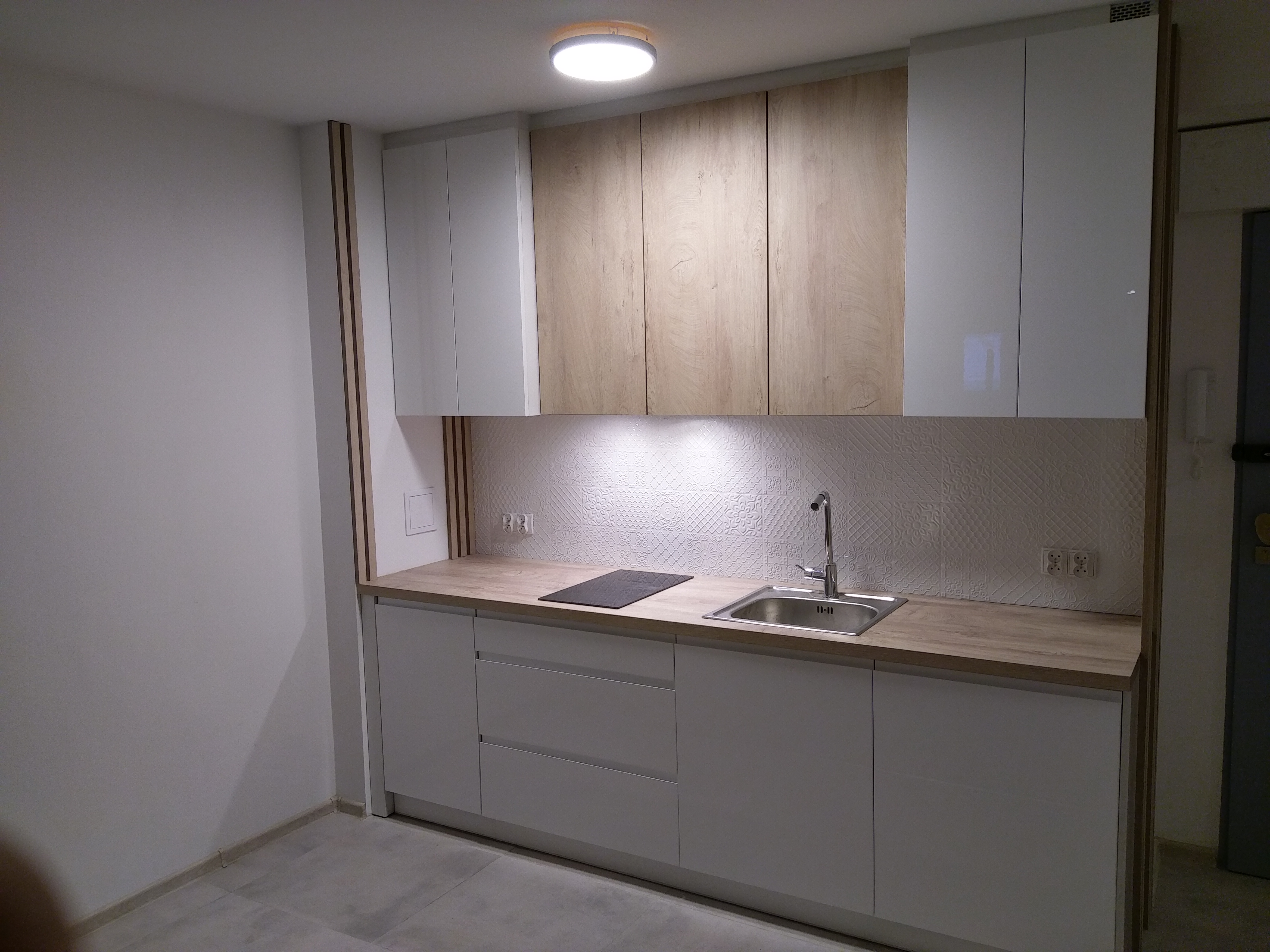 Nowoczesna kuchnia z białymi szafkami dolnymi i górnymi, oraz drewnopodobnymi szafkami górnymi, oświetlona lampą sufitową i listwą LED pod szafkami, z płytkami dekoracyjnymi na ścianie.