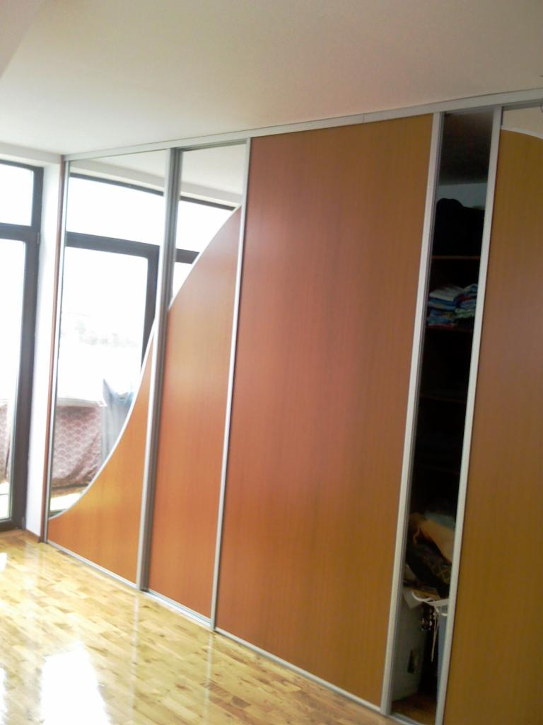 Brązowa zabudowa wnęki z przesuwnymi drzwiami, częściowo lustrzanymi, z widoczną zawartością półek w otwartej części, na parkiecie.