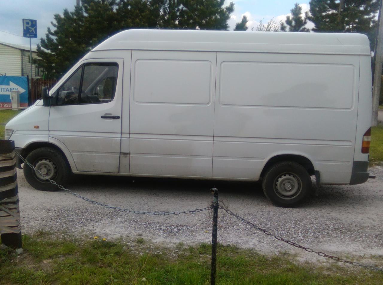 Biały, brudny furgon dostawczy zaparkowany na żwirowej drodze, widoczny z boku, z łańcuchem w pierwszym planie i budynkami w tle.