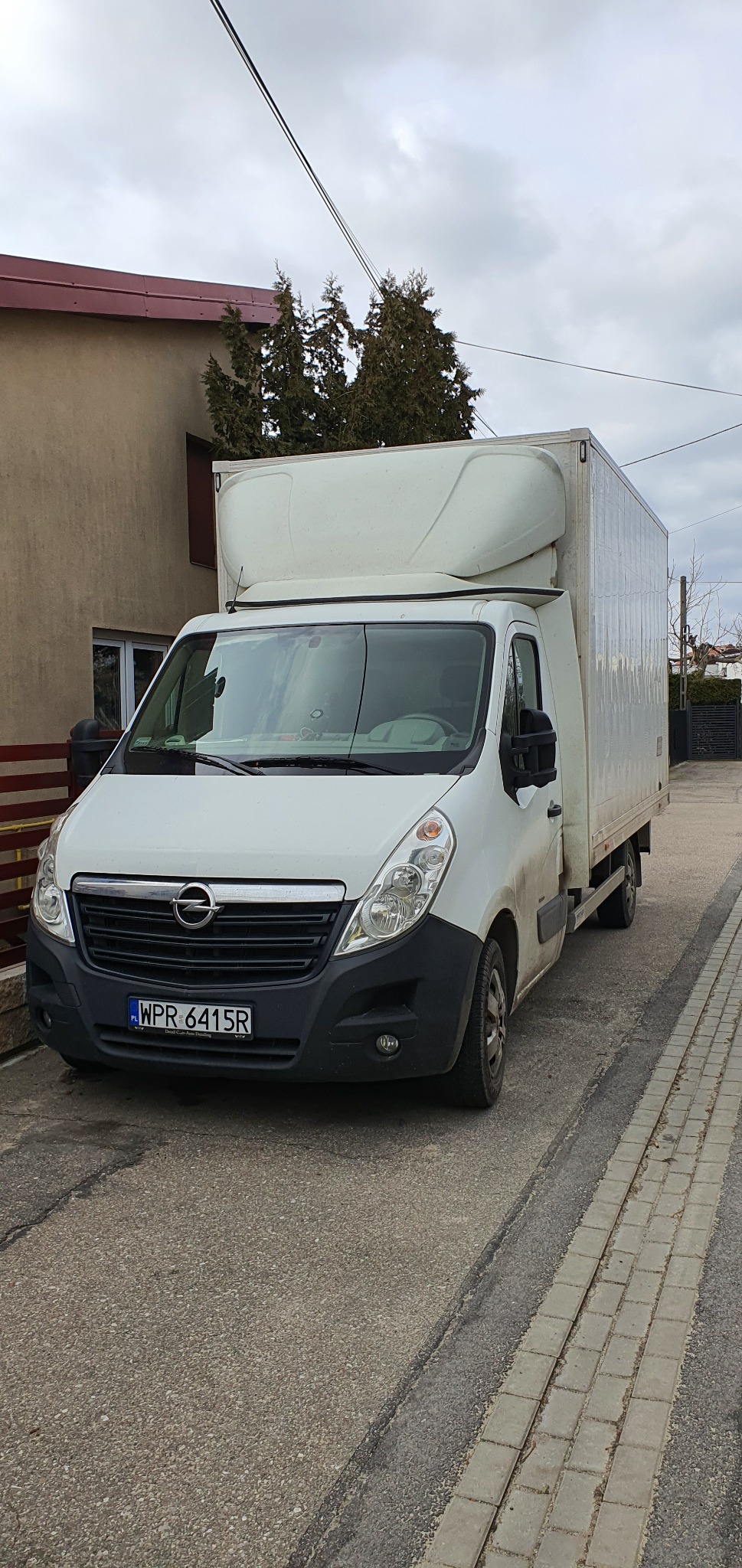 Biały samochód dostawczy Opel Master z zabudową kontenerową, zaparkowany na ulicy z kostki brukowej przed budynkiem.