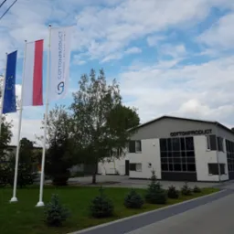 Budynek firmy Cotton Product z trzema masztami flagowymi z flagami Unii Europejskiej, Polski i logo firmy na tle błękitnego nieba z chmurami.