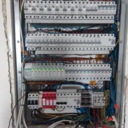 Rozdzielnica elektryczna z widocznymi bezpiecznikami, przewodami i elementami zabezpieczeń, zamontowana w ścianie z surowym wykończeniem.