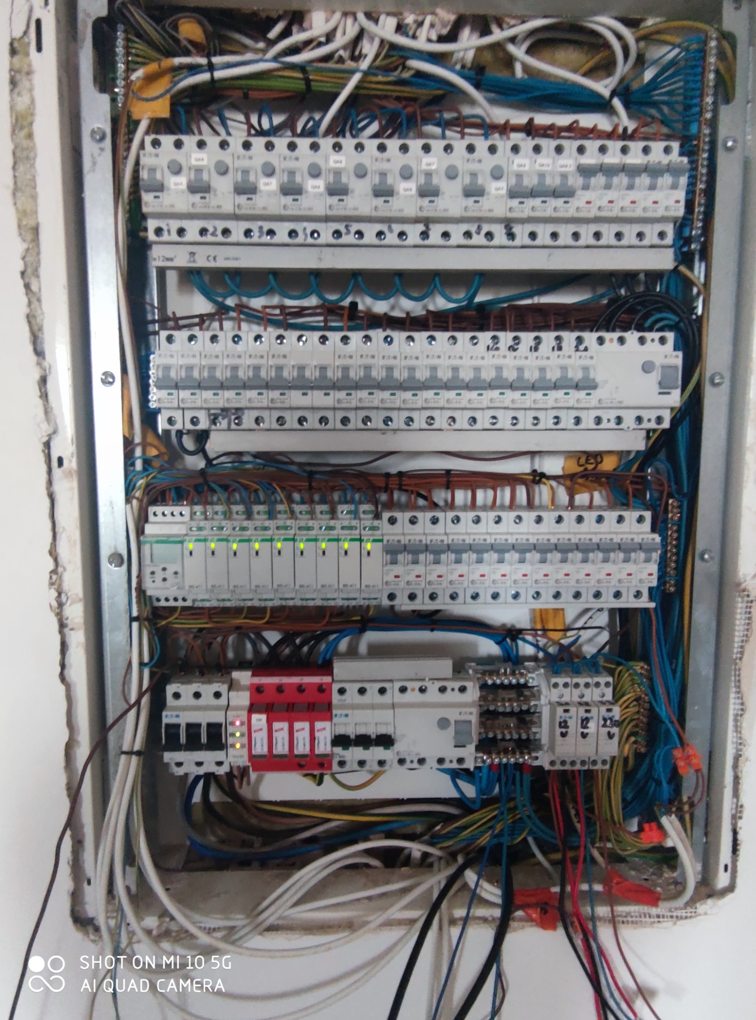 Rozdzielnica elektryczna z widocznymi bezpiecznikami, przewodami i elementami zabezpieczeń, zamontowana w ścianie z surowym wykończeniem.