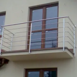 Balkon z nowoczesną, poziomą balustradą ze stali nierdzewnej, zamontowaną na beżowej elewacji budynku z brązowymi oknami. Widoczny fragment dachu z brązową dachówką.