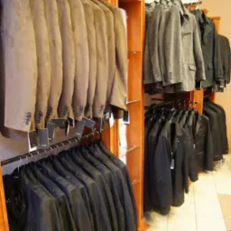 Rzędy garniturów w odcieniach brązu, szarości i czerni wiszą na wieszakach w sklepie odzieżowym, z drewnianymi regałami i jasną podłogą w kratkę.