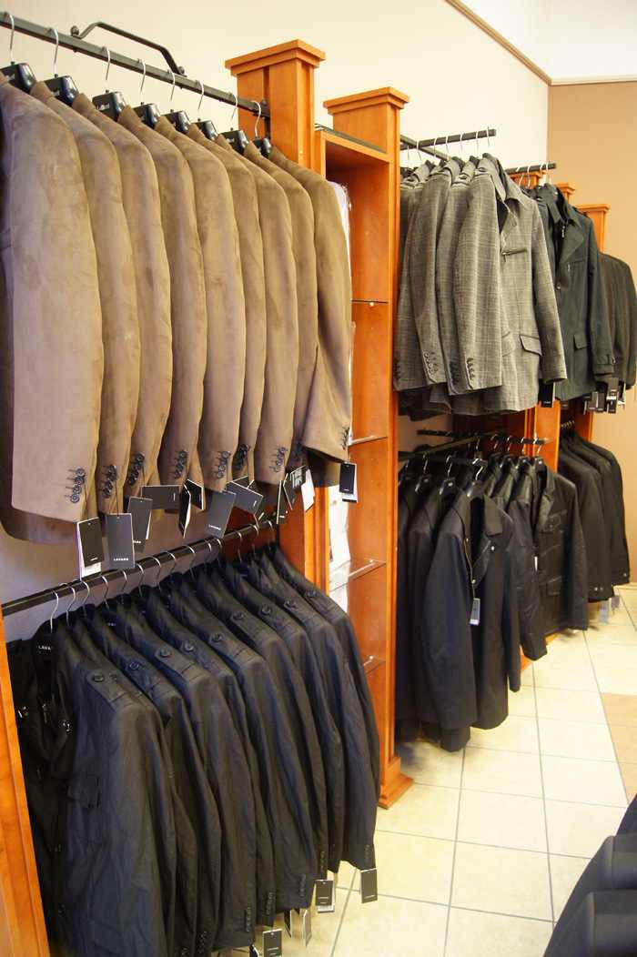 Rzędy garniturów w odcieniach brązu, szarości i czerni wiszą na wieszakach w sklepie odzieżowym, z drewnianymi regałami i jasną podłogą w kratkę.