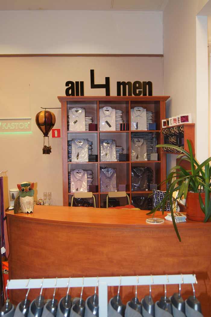 Wnętrze sklepu odzieżowego z drewnianą ladą, regałem wypełnionym zapakowanymi koszulami męskimi, dekoracyjnym balonem na ogrzane powietrze i napisem 'all 4 men' na ścianie.