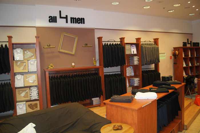 Wnętrze salonu z męską odzieżą wizytową: rzędy garniturów na wieszakach, koszule w pudełkach, drewniane regały i lada z ubraniami, w tle kanapa i logo sklepu 'all 4 men'.