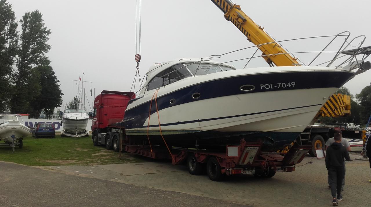 Jacht motorowy z numerem rejestracyjnym POL-7049 transportowany na niskopodwoziowej naczepie, podnoszony przez dźwig w pobliżu innych łodzi i drzew.
