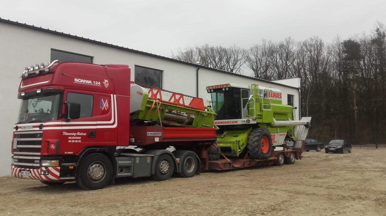 Czerwona ciężarówka Scania przewozi na niskopodwoziowej naczepie kombajn zbożowy marki Claas Dominator, widoczne budynki w tle.