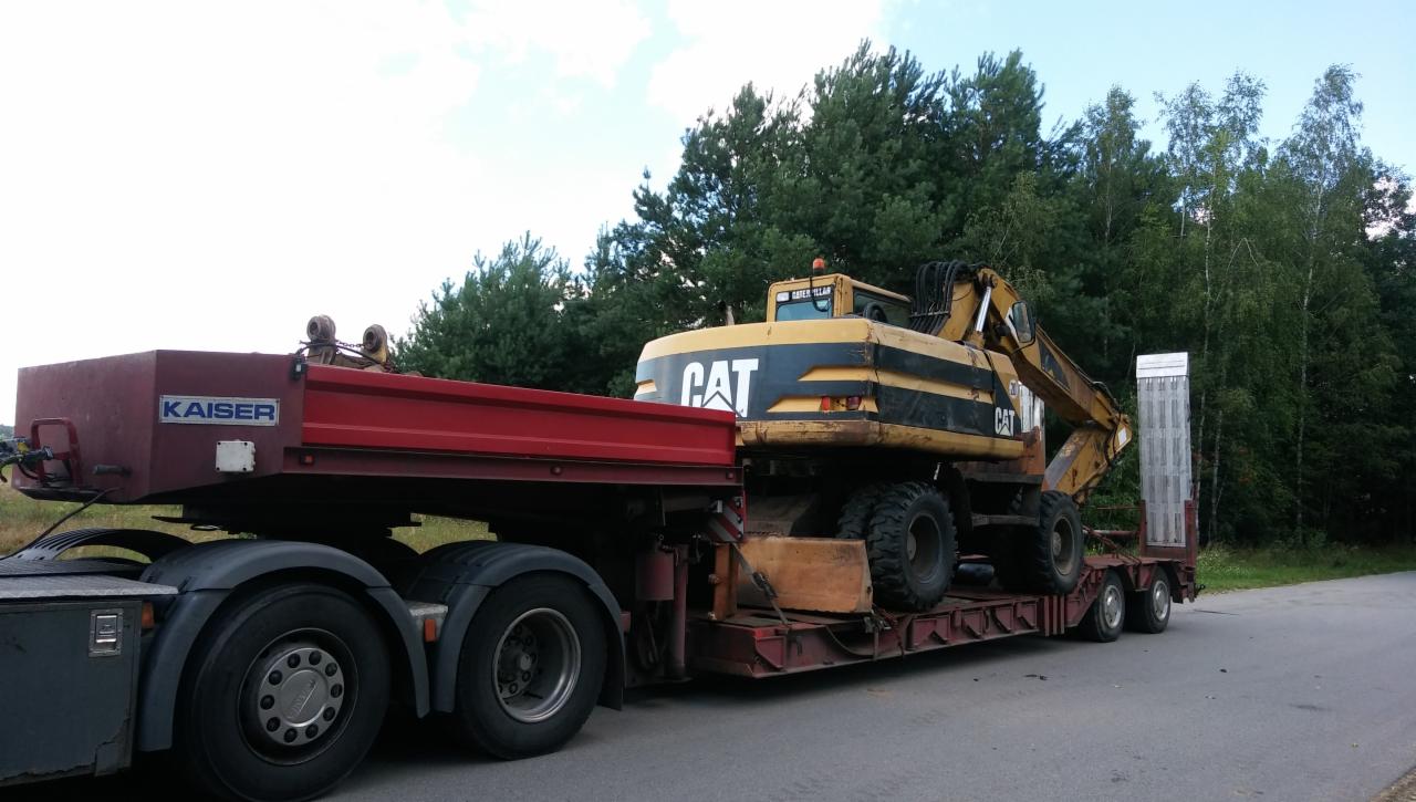 Ciężka koparka marki CAT przewożona na niskopodwoziowej naczepie przez firmę z Wołomina, na tle zieleni.
