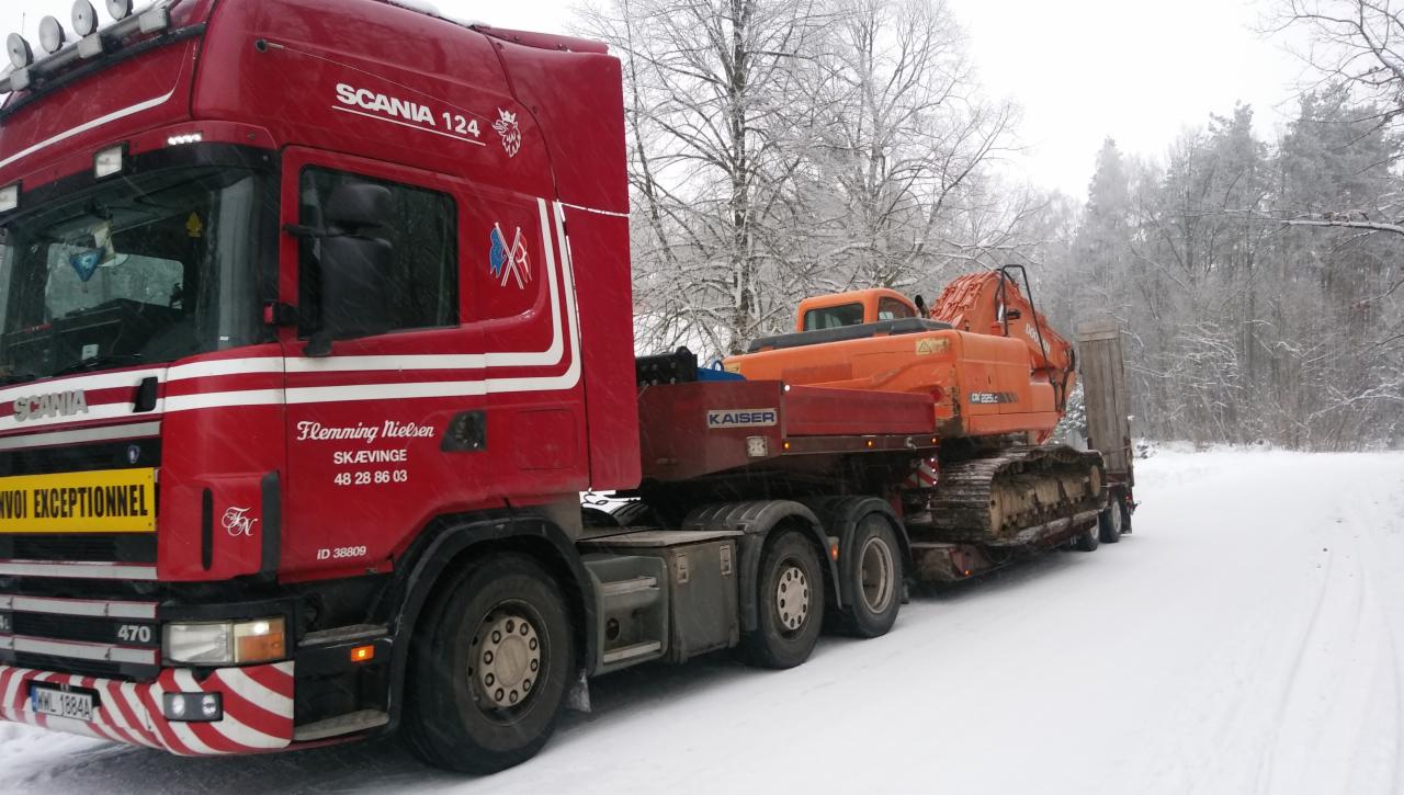Czerwona ciężarówka Scania 124 z pomarańczową koparką na naczepie Kaiser, transportowana po ośnieżonej drodze w zimowy dzień.