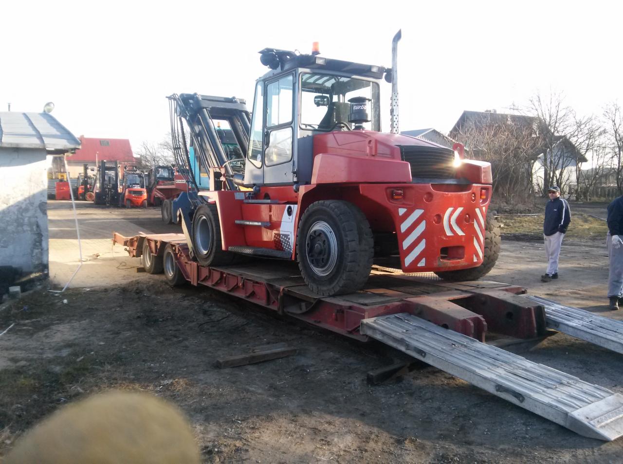 Czerwony wózek widłowy specjalnego przeznaczenia z białymi pasami na lawecie, w tle budynki gospodarcze i inne wózki widłowe, widoczni dwaj mężczyźni.