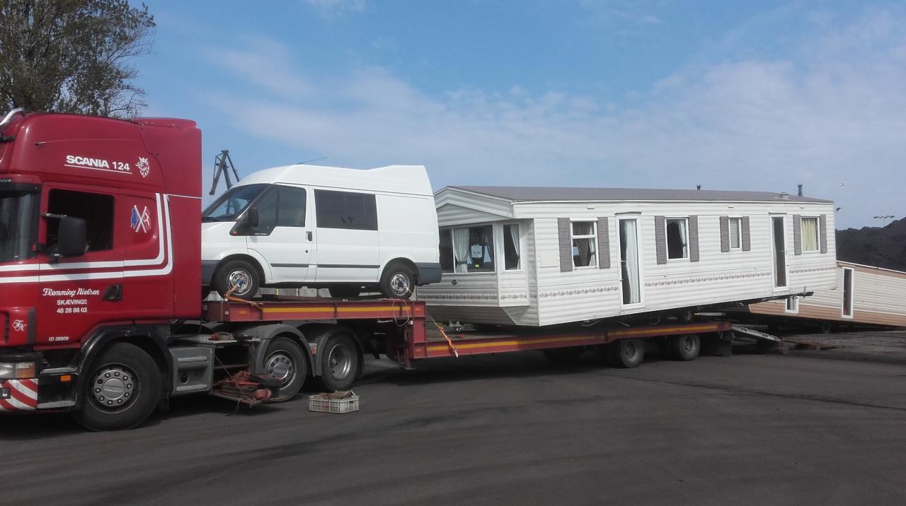 Czerwona ciężarówka Scania przewozi na lawecie biały domek holenderski i białego busa na tle błękitnego nieba.