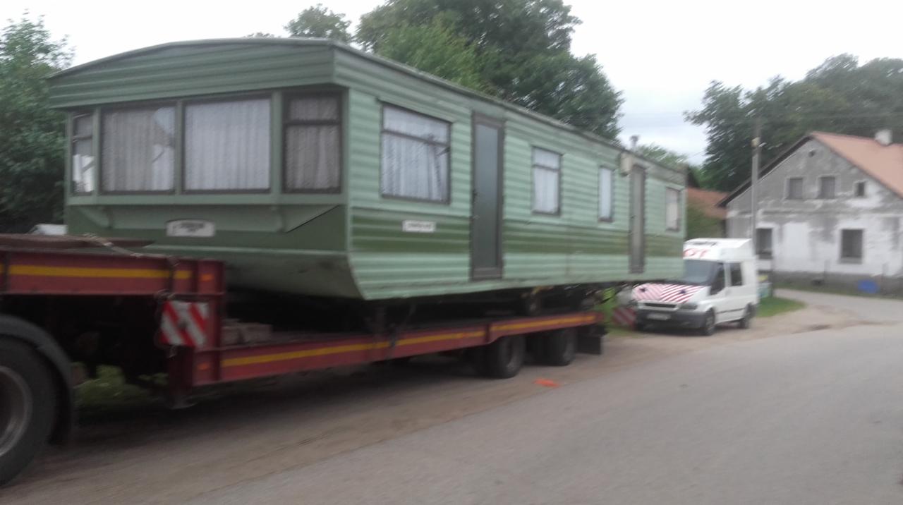Zielony domek holenderski o wymiarach 11,5m x 3,7m transportowany na niskopodwoziowej naczepie ciężarówki, z białym busem eskortującym w tle.