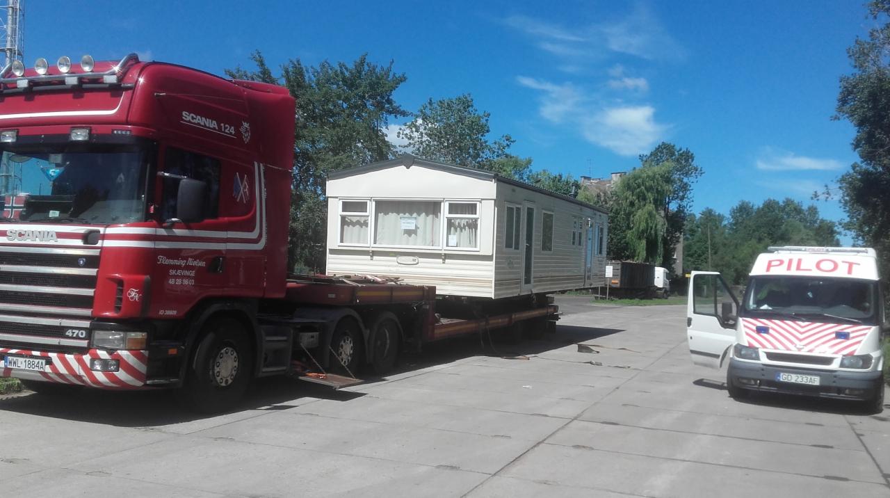 Czerwona ciężarówka Scania 124 przewozi biały domek holenderski na niskopodwoziowej platformie, z białym busem pilotującym z otwartymi drzwiami po prawej stronie, na tle drzew i błękitnego nieba.