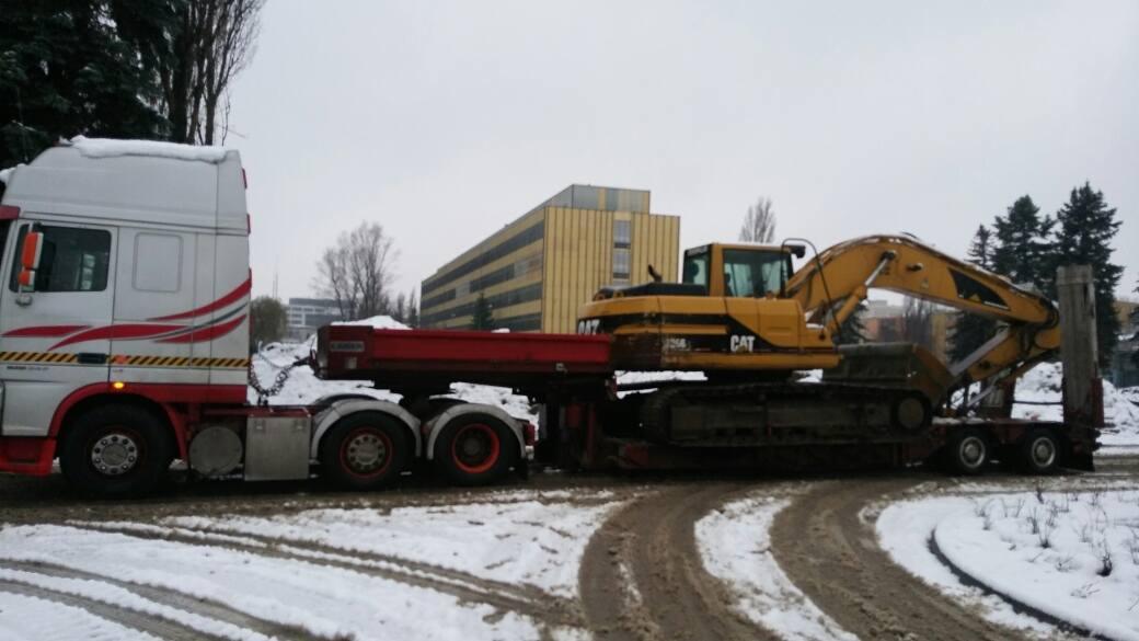 Ciężarówka z naczepą niskopodwoziową przewozi żółtą koparkę CAT na tle budynku w zimowej scenerii.