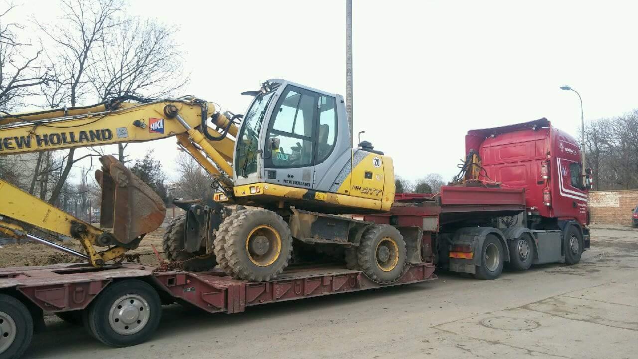 Żółto-szara koparka New Holland na niskopodwoziowej naczepie ciągniętej przez czerwony ciągnik siodłowy Scania, widok z boku na placu.
