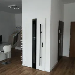 Nowoczesne drzwi zewnętrzne w kolorze białym z czarnymi akcentami i długim, pionowym uchwytem, prezentowane w jasnym wnętrzu salonu sprzedaży z ekspozycją paneli podłogowych w tle.