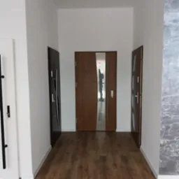 Korytarz z trzema parami drzwi: białe drzwi wewnętrzne z czarną klamką po lewej, brązowe drzwi wewnętrzne z pionowym przeszkleniem na wprost i brązowe drzwi wewnętrzne po prawej, na podłodze panele...