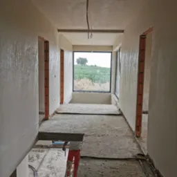 Wnętrze w trakcie prac wykończeniowych: świeżo otynkowane ściany w korytarzu z widokiem na pole, pracownik na drabinie w czerwonych spodniach, widoczne przewody elektryczne na podłodze.